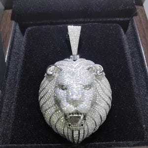 May include: A silver-colored lion pendant encrusted with small, sparkling stones. The pendant is in a black box, showcasing the detailed craftsmanship of the lion's face, mane, and open mouth. The pendant has a rectangular bail.