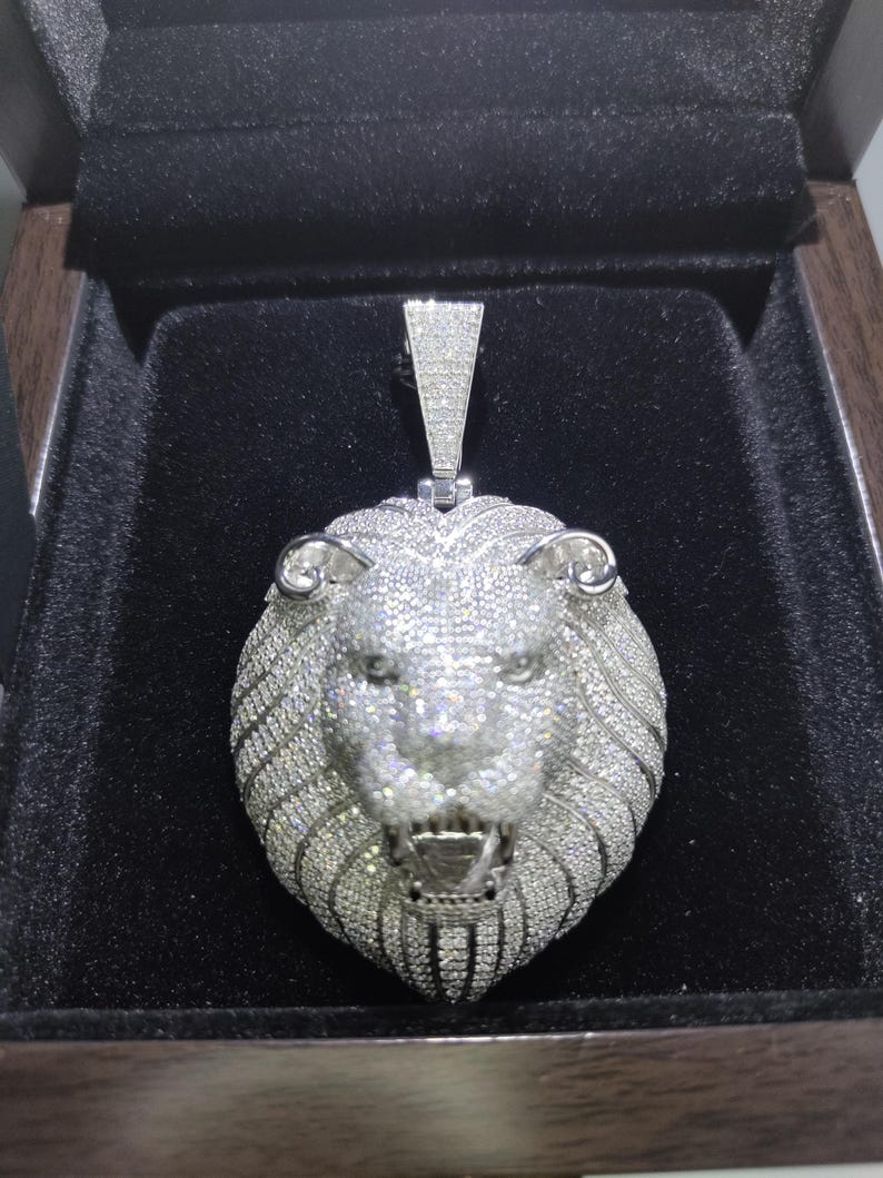 May include: A silver-colored lion pendant encrusted with small, sparkling stones. The pendant is in a black box, showcasing the detailed craftsmanship of the lion's face, mane, and open mouth. The pendant has a rectangular bail.