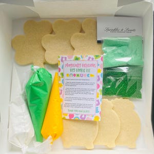 May include: A DIY cookie kit in a white box. The kit includes clover-shaped cookies, green and yellow icing bags, green sprinkles, and a card that reads "Magically Delicious DIY Cookie Kit". The kit is from "Sprinkles & Sweets".