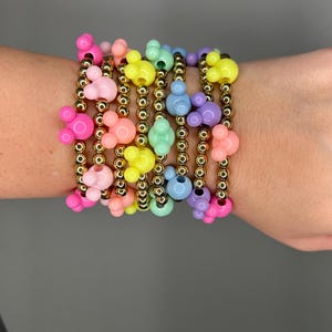 Puede incluir: Una pila de pulseras coloridas con cuentas doradas y dijes de cabeza de Mickey Mouse. Las pulseras son en tonos de rosa, amarillo, azul y verde.