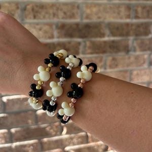 Puede incluir: Tres pulseras de cuentas con cuentas en forma de Mickey Mouse en negro, blanco y dorado. Las pulseras son el&aacute;sticas y se muestran en una mu&ntilde;eca. El fondo es una pared de ladrillos. Estas pulseras son un accesorio divertido.