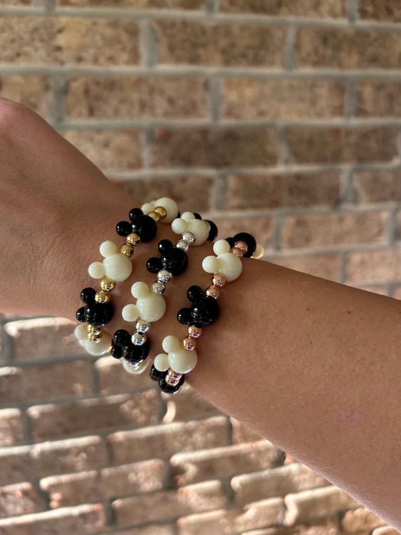Puede incluir: Tres pulseras de cuentas con cuentas en forma de Mickey Mouse en negro, blanco y dorado. Las pulseras son el&aacute;sticas y se muestran en una mu&ntilde;eca. El fondo es una pared de ladrillos. Estas pulseras son un accesorio divertido.