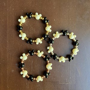 Puede incluir: Tres pulseras de cuentas con un patr&oacute;n repetido de siluetas de Mickey Mouse negras y crema. Las pulseras tienen cuentas doradas, plateadas y cobrizas. Las pulseras se exhiben sobre una superficie marr&oacute;n.