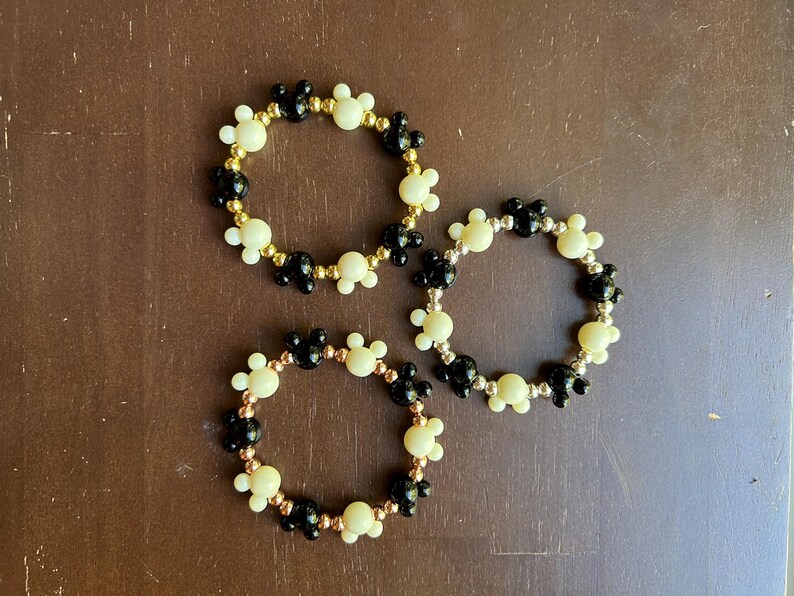 Puede incluir: Tres pulseras de cuentas con un patr&oacute;n repetido de siluetas de Mickey Mouse negras y crema. Las pulseras tienen cuentas doradas, plateadas y cobrizas. Las pulseras se exhiben sobre una superficie marr&oacute;n.