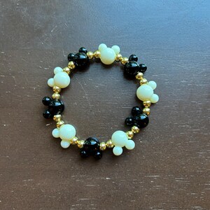 Puede incluir: Una pulsera de cuentas con formas de cabeza de Mickey Mouse negras y crema intercaladas con cuentas doradas. La pulsera es circular y descansa sobre una superficie marr&oacute;n.