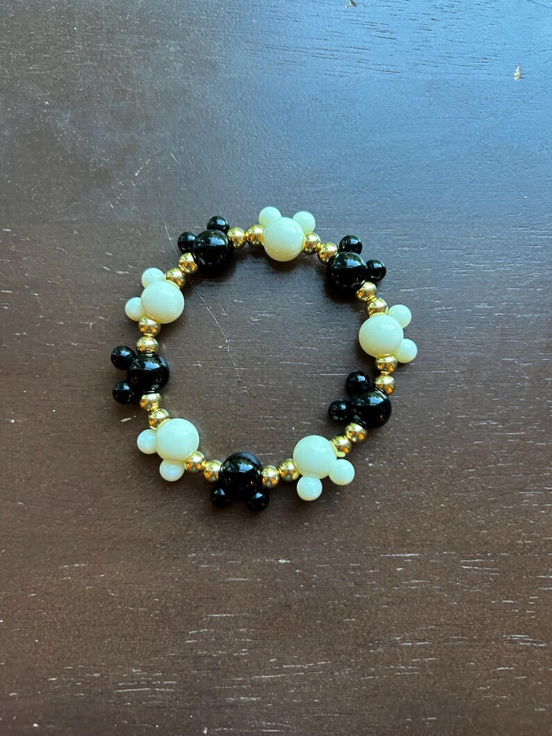 Puede incluir: Una pulsera de cuentas con formas de cabeza de Mickey Mouse negras y crema intercaladas con cuentas doradas. La pulsera es circular y descansa sobre una superficie marr&oacute;n.