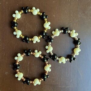 Puede incluir: Tres pulseras de cuentas. Cada pulsera presenta un patr&oacute;n repetido de cuentas negras y crema con forma de Mickey Mouse. Las cuentas est&aacute;n ensartadas en un alambre dorado. Las pulseras se exhiben sobre una superficie marr&oacute;n.