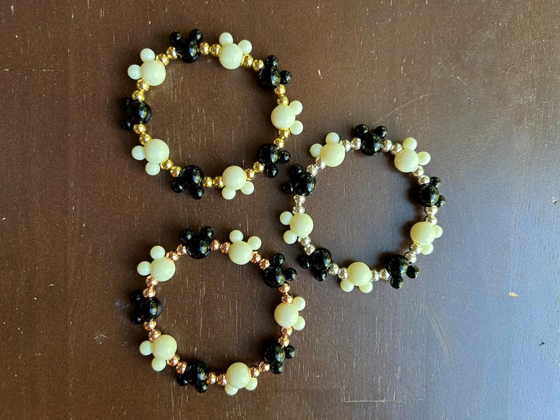Puede incluir: Tres pulseras de cuentas. Cada pulsera presenta un patr&oacute;n repetido de cuentas negras y crema con forma de Mickey Mouse. Las cuentas est&aacute;n ensartadas en un alambre dorado. Las pulseras se exhiben sobre una superficie marr&oacute;n.