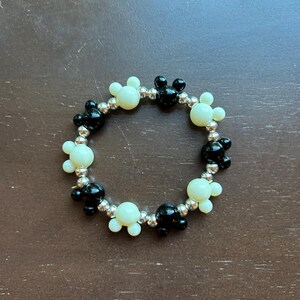 Puede incluir: Una pulsera de cuentas con cuentas en forma de Mickey Mouse negras y blanquecinas alternadas. Las cuentas est&aacute;n ensartadas en una banda el&aacute;stica plateada. La pulsera es circular y se asienta sobre una superficie marr&oacute;n.