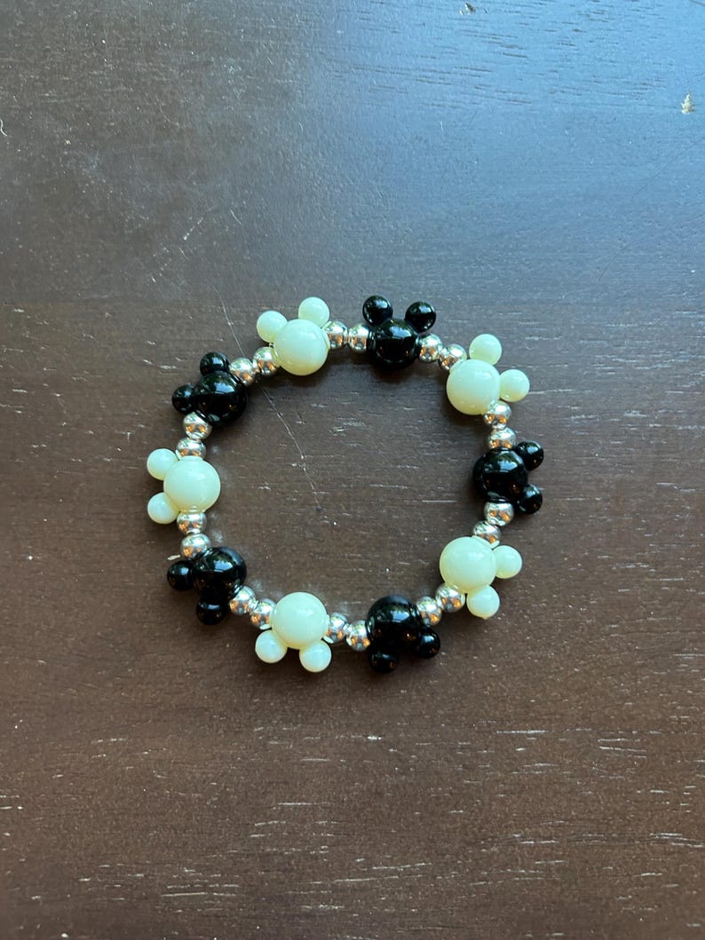 Puede incluir: Una pulsera de cuentas con cuentas en forma de Mickey Mouse negras y blanquecinas alternadas. Las cuentas est&aacute;n ensartadas en una banda el&aacute;stica plateada. La pulsera es circular y se asienta sobre una superficie marr&oacute;n.