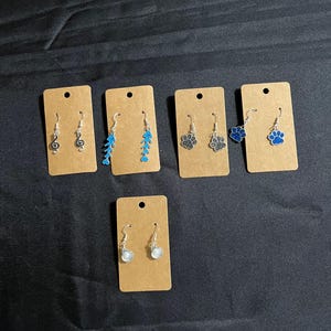 May include: A collection of handmade earrings displayed on brown cards. The earrings feature various designs, including musical notes, blue fish bones, paw prints, and seashells. The earrings are silver-toned and have a simple hook design.
