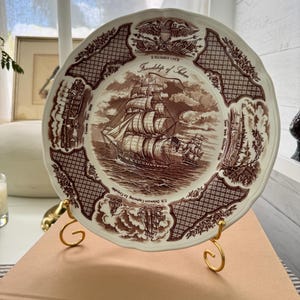 May include: A decorative plate with a brown and white nautical design. The plate features a detailed illustration of a tall ship sailing on the ocean, surrounded by decorative borders and text. The plate is displayed on a gold-colored stand.