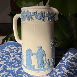 May include: Cream-colored ceramic pitcher with blue bas-relief designs. The pitcher features a handle and spout, with a band of grapevines around the top. The body of the pitcher depicts classical figures and a tree. A decorative piece for the home.