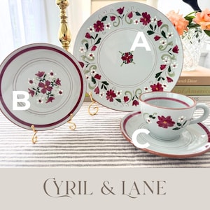 May include: A light blue floral-patterned dinnerware set. The set includes a large plate, a smaller plate, and a teacup with a saucer. Each piece features a burgundy rim and floral design. The brand name "CYRIL & LANE" is at the bottom.