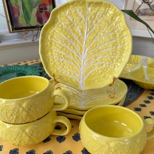 May include: A collection of yellow ceramic tableware, including plates and cups, with a cabbage leaf design. The plates are supported by gold-colored stands. The cups are stacked and have handles. The set is arranged on a yellow tablecloth.