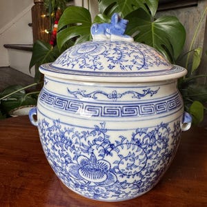 May include: A white and blue ceramic lidded jar with a floral and geometric design. The jar has two small handles and a blue figurine on top of the lid. The jar is sitting on a wooden surface.