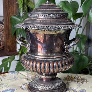 May include: A silver-plated antique urn with a lid, handles, and a decorative base. The urn has a fluted lower section with a copper-colored band. The lid is topped with a small finial. The urn is displayed on a floral patterned tablecloth.