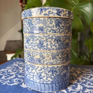 May include: A stacked ceramic container with a lid, featuring a blue and white floral pattern. The container is composed of several sections, each adorned with detailed designs. The piece is set on a blue patterned cloth, showcasing its decorative nature.