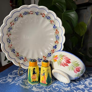 May include: A white ceramic serving bowl with a scalloped edge and a blue, red, and green floral design. Two yellow salt and pepper shakers with painted figures and a matching soap dish are also present.