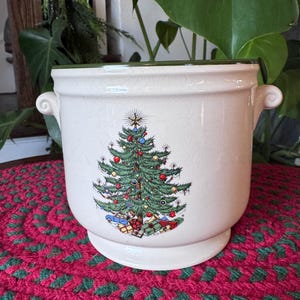 May include: Cream-colored ceramic planter with a Christmas tree design. The tree is decorated with ornaments and presents. The planter has two handles and a pedestal base. The rim is trimmed with green.