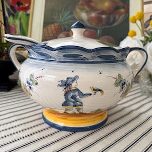 May include: A white ceramic soup tureen with a lid and ladle, featuring a blue wavy rim and handles. The tureen is decorated with a painted scene of a person with a bird, and floral accents. The base is yellow.