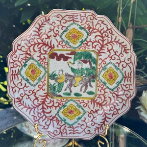 May include: An octagonal, antique ceramic plate with a detailed Asian-inspired design. The plate features a central square scene with figures on horseback and a tree, surrounded by red and green decorative patterns and yellow accents.