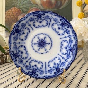 May include: A white and blue antique plate with a scalloped edge, displayed on a gold stand. The plate features a detailed blue floral pattern and a central starburst design. The plate is a decorative item.