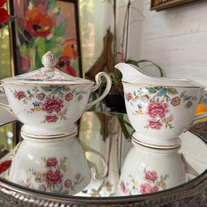 May include: A white porcelain sugar bowl with a lid and a creamer. Both pieces are decorated with a floral pattern in shades of red, pink, blue, and green, with gold trim. The set is displayed on a mirrored surface.