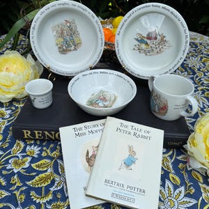 May include: A collection of Peter Rabbit themed items, including two white plates, a bowl, a mug, and a small cup, all with illustrations. Two Beatrix Potter books are also present, along with a dark book and a floral tablecloth.