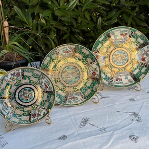 May include: Three decorative ceramic plates with intricate designs in green, gold, and red, displayed on gold-colored stands. The plates feature detailed patterns and are arranged on a white tablecloth.