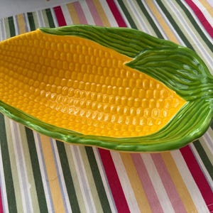 May include: A ceramic serving dish shaped like an ear of corn with a bright yellow textured kernel center and glossy green husk leaves wrapping around the edges. The elongated oval bowl features a realistic embossed pattern of corn kernels.