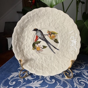May include: Cream-colored decorative plate with a bird and floral design. The plate features a detailed embossed floral pattern around the rim and a painted image of a bird with long tail feathers. The plate is displayed on a gold-colored metal stand.