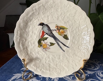 May include: Cream-colored decorative plate with a bird and floral design. The plate features a detailed embossed floral pattern around the rim and a painted image of a bird with long tail feathers. The plate is displayed on a gold-colored metal stand.
