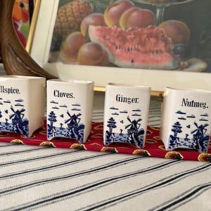May include: Four white ceramic spice jars labeled Allspice, Cloves, Ginger, and Nutmeg. Each jar features a blue and white windmill design. The jars are displayed on a red patterned runner.