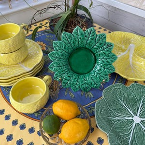 May include: A collection of decorative ceramic tableware, including yellow teacups and saucers, leaf-shaped plates, and a green floral-shaped plate. Two lemons and a lime sit in a glass dish. The items are arranged on a yellow tablecloth with a blue floral pattern.