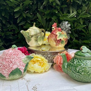 May include: Four decorative teapots in various designs and colors. One is pink with a floral pattern, another is green with a lattice design, and a third is yellow with a rooster design. The teapots are on a white tablecloth.