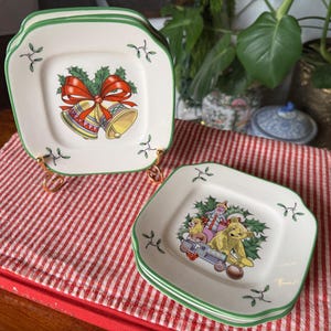 May include: Set of square ceramic Christmas plates with a green border. One plate features a red bow and golden bells, while another displays a Santa Claus and teddy bear design. The plates rest on a red and white striped cloth.