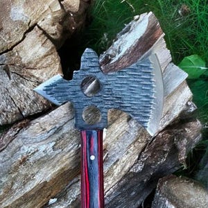 May include: A handcrafted axe with a textured gray steel head and a red, black, and white striped wooden handle. The axe head features a blade, a saw-like edge, and two circular cutouts. The handle is secured with metal hardware.