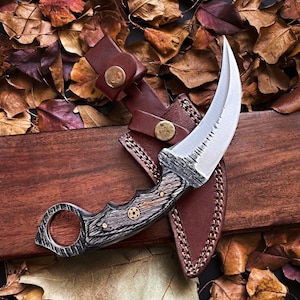 May include: A curved karambit knife with a wooden handle and a silver blade, resting in a brown leather sheath. The handle features brass accents. The knife is displayed on a wooden surface with autumn leaves.