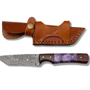 May include: A Damascus steel knife with a dark wood handle and a purple resin inlay. The knife is paired with a brown leather sheath. The blade has a unique pattern, and the handle is secured with brass rivets.