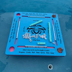 May include: A light blue square floating game board for Pool Jong with a central black lotus graphic and text. The board features four slots for tiles, printed rules regarding Jokers and Dragons, and four circular corner cutouts for drinks.
