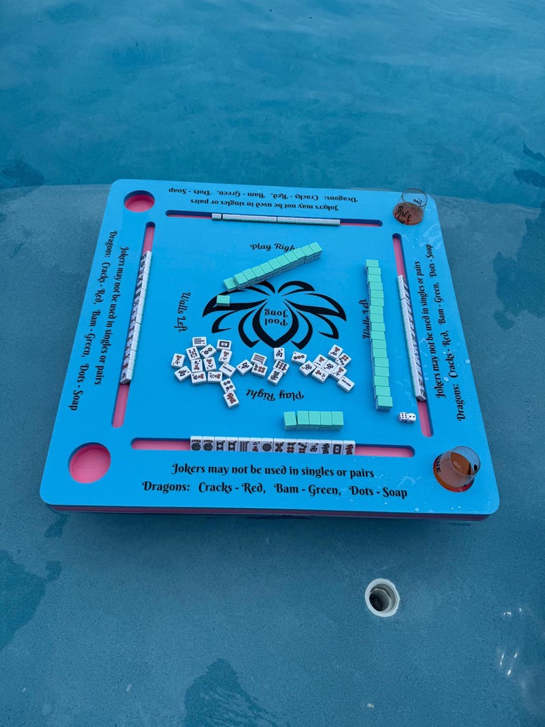 May include: A light blue square floating game board for Pool Jong with a central black lotus graphic and text. The board features four slots for tiles, printed rules regarding Jokers and Dragons, and four circular corner cutouts for drinks.