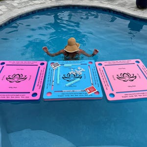 May include: Three square floating game boards for a tile-based game, two pink and one blue, each featuring a black lotus flower graphic in the center. The boards have slots for tiles and printed text instructions, with game tiles arranged on the blue board.