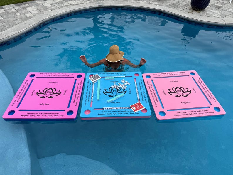 May include: Three square floating game boards for a tile-based game, two pink and one blue, each featuring a black lotus flower graphic in the center. The boards have slots for tiles and printed text instructions, with game tiles arranged on the blue board.