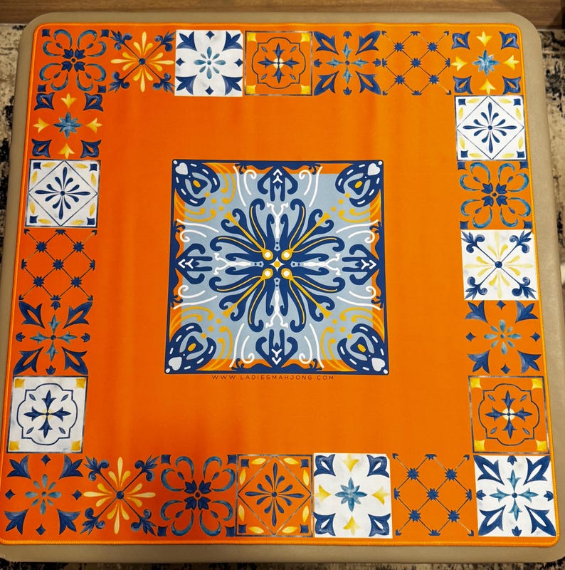 K&ouml;nnte beinhalten: Quadratischer, orangefarbener Mahjong-Tisch mit dekorativem Fliesendesign. Die Mitte zeigt ein gro&szlig;es blau-wei&szlig;es Blumenmuster, und der Rand hat kleinere Fliesenmotive in Blau, Wei&szlig; und Orange. Die Website www.ladiesmahjong.com ist sichtbar.