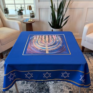 May include: A square blue tablecloth featuring a colorful abstract painting of a menorah in a gold frame. The border has gold stripes and Star of David symbols. The tablecloth is set on a table with white legs.