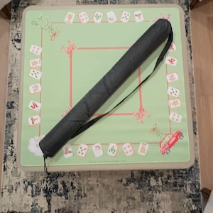 May include: A green Mahjong game mat with a red border and decorative elements. A black carrying case with a shoulder strap rests on the mat. The mat features various game tiles and festive designs.