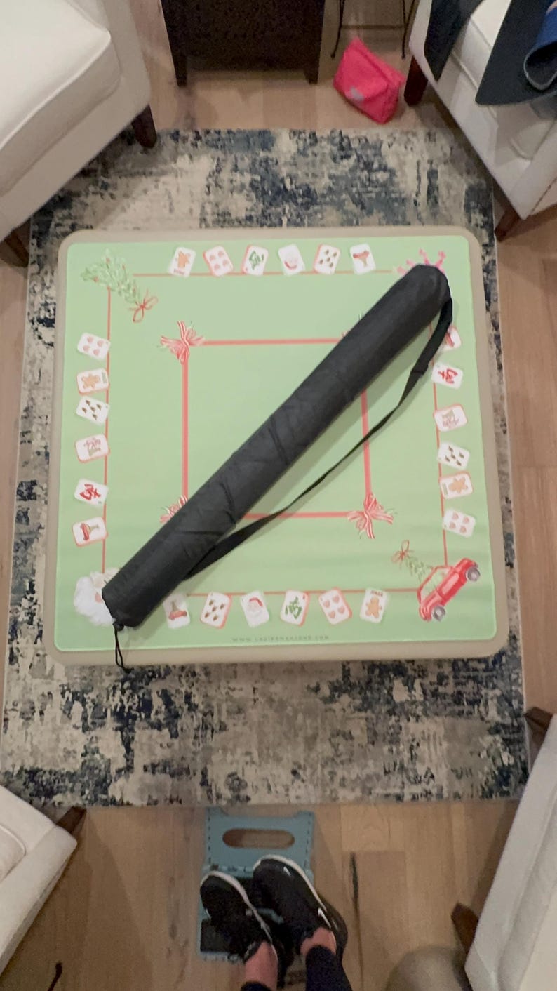 May include: A green Mahjong game mat with a red border and decorative elements. A black carrying case with a shoulder strap rests on the mat. The mat features various game tiles and festive designs.