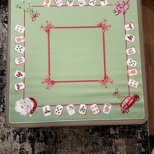 May include: A green Mahjong table cover with a red border and bow details. The cover displays Christmas-themed card illustrations, a Santa Claus graphic, and a red car with a Christmas tree. The website address www.ladiesmahjong.com is printed on the cover.