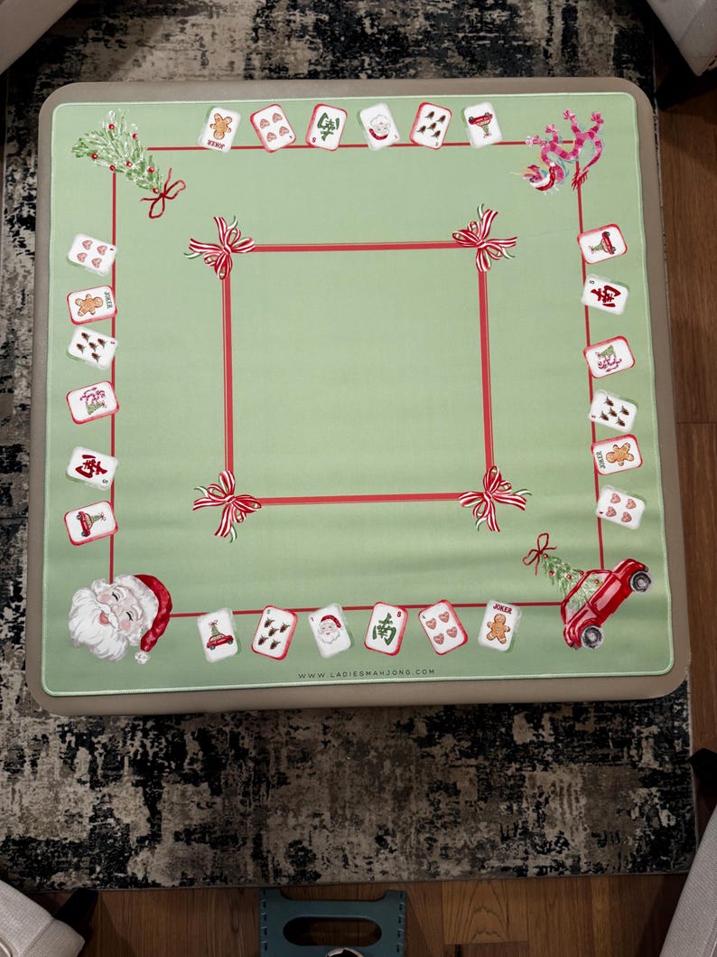 May include: A green Mahjong table cover with a red border and bow details. The cover displays Christmas-themed card illustrations, a Santa Claus graphic, and a red car with a Christmas tree. The website address www.ladiesmahjong.com is printed on the cover.